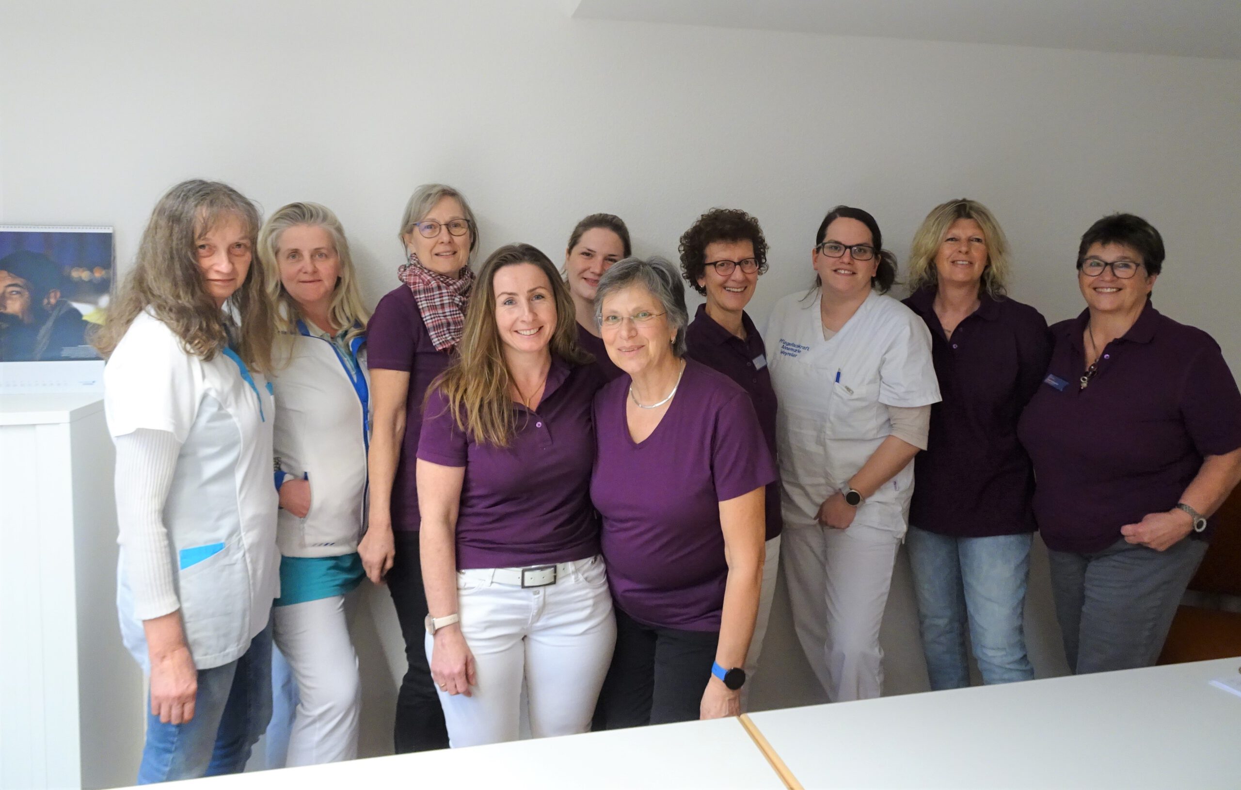 Eine Gruppe von zehn Frauen, einige in lila, andere in weiß gekleidet, steht lächelnd in einem Raum.