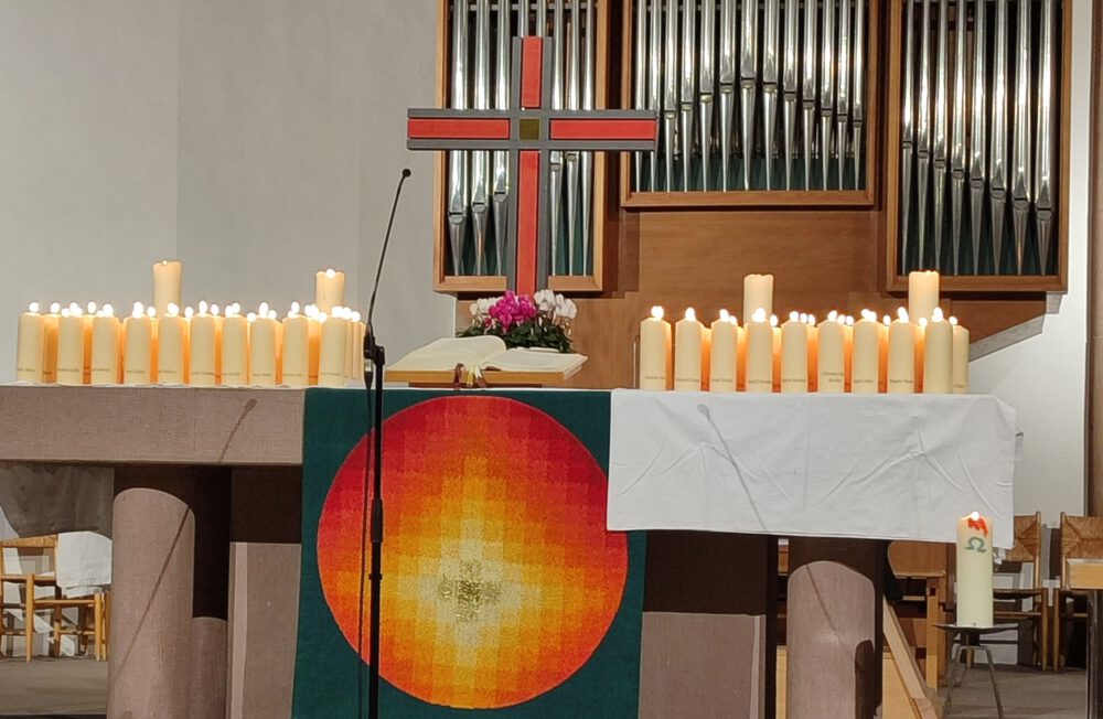 Ein mit vielen brennenden Kerzen, einem großen roten Kreuz, einem Banner mit einem roten und gelben Kreis und rosa Blumen geschmückter Altar, mit Orgelpfeifen im Hintergrund.