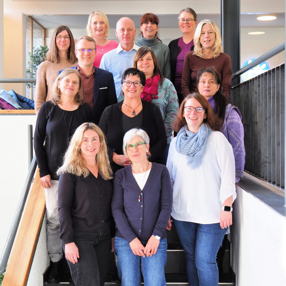 Eine Gruppe von fünfzehn Erwachsenen, sowohl Männer als auch Frauen, stehen und sitzen auf einer Treppe in einem Innenraum und lächeln in die Kamera. Sie sind leger gekleidet und sitzen in zwei Reihen bei hellem Tageslicht.